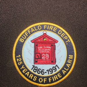 May include: A round, gold-trimmed patch with a blue and white background. The patch features a red fire alarm box with the number 29 on it. The text "BUFFALO FIRE DEPT. 125 YEARS OF FIRE ALARM 1866-1991" is written around the edge of the patch.