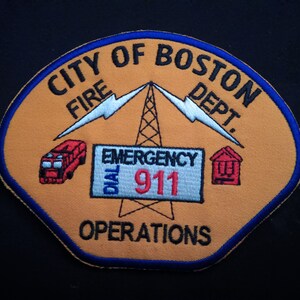 Puede incluir: Un parche bordado en amarillo y azul con el texto "City of Boston Fire Dept." y "Emergency Dial 911 Operations". El parche presenta un camión de bomberos, una torre de radio con rayos y una casa.