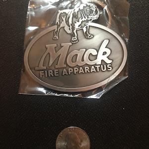 May include: A silver-colored oval keychain featuring a bulldog illustration above the words "Mack FIRE APPARATUS." The keychain is in a clear plastic wrapper, with a U.S. quarter for size comparison.