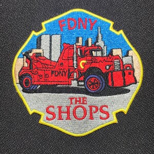 Könnte beinhalten: Ein gestickter Aufnäher in Rot und Gelb mit dem Text "FDNY The Shops" und einem Cartoon-Bild eines roten Feuerwehrautos mit dem Text "FDNY" an der Seite. Das Auto fährt durch eine Skyline.