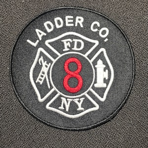 Könnte beinhalten: Schwarzer und weißer gestickter Aufnäher mit einer roten Nummer 8 in der Mitte. Der Aufnäher zeigt ein Feuerwehrsymbol mit dem Text "FD NY" und "LADDER CO."