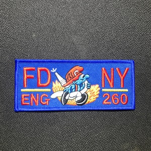Könnte beinhalten: Blauer und roter gestickter Aufnäher mit dem Text "FDNY ENG 260" und einem Comic-Feuerwehrmann in rotem Helm und blauer Latzhose, der einen Feuerwehrschlauch hält.