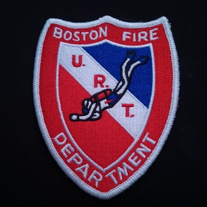 Può includere: Una toppa ricamata in rosso, bianco e blu con le parole "BOSTON FIRE DEPARTMENT" attorno al bordo. Un subacqueo è raffigurato al centro con le lettere "U.R.T." sopra.