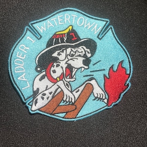 Op de afbeelding: Een geborduurde patch in blauw en wit met een cartoon dalmatiër die een brandweerhelm draagt en een ladder vasthoudt. De patch heeft de tekst "Ladder 1 Watertown" rond de rand.
