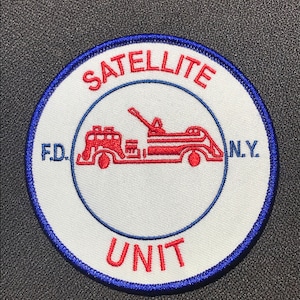 Könnte beinhalten: Ein weißer runder Patch mit einem blauen Rand. Der Patch hat roten Text, der "SATELLITE UNIT" und "F.D.N.Y." lautet. Ein rotes und weißes Feuerwehrauto befindet sich in der Mitte des Patches.