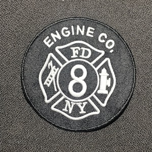 Könnte beinhalten: Ein schwarz-weißer, gestickter Aufnäher mit dem Text "ENGINE CO." oben und "FD" in der Mitte. Die Zahl "8" befindet sich in der Mitte, mit "NY" unten. Der Aufnäher zeigt ein Malteserkreuz-Design.