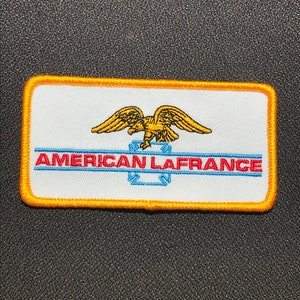 Könnte beinhalten: Ein weißer gestickter Aufnäher mit gelbem Rand. Der Aufnäher zeigt einen gelben Adler mit roten Akzenten und den Text "AMERICAN LAFRANCE" in Blau und Rot.