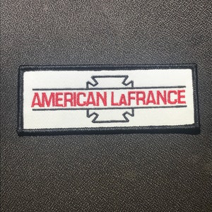 Könnte beinhalten: Rechteckiger Aufnäher mit dem Text "AMERICAN LAFRANCE" in roten Buchstaben auf weißem Grund. Der Aufnäher hat einen schwarzen Rand und eine schwarze Grafik über dem Text. Der Hintergrund ist eine strukturierte, dunkelgraue Oberfläche.