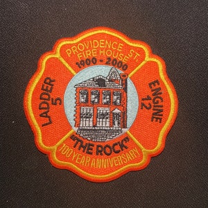 May include: Orange and yellow embroidered patch with the text "Providence St. Fire House 1900-2000" and "Ladder 5 Engine 212" around a building illustration. The text "The Rock 100 Year Anniversary" is also included.