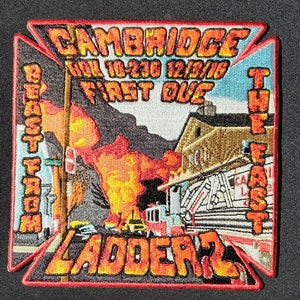 Può includere: Una toppa ricamata in rosso, bianco e blu con le parole "CAMBRIDGE" e "LADDER 2" in un carattere arancione in grassetto. La toppa presenta anche le parole "FIRST DUE" e "THE EAST" in un carattere più piccolo. Lo sfondo della toppa rappresenta una scena di un edificio in fiamme con i vigili del fuoco che intervengono.