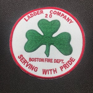 Op de afbeelding: Een witte en rode geborduurde patch met een groene klaver in het midden. De tekst rond de klaver luidt "Ladder 20 Company Serving With Pride Boston Fire Dept."
