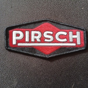 May include: Embroidered patch featuring the word "PIRSCH" in white, with a silver outline, set against a red backdrop. The patch has a black border and a distinctive hexagonal form. Likely designed for application on clothing or bags.