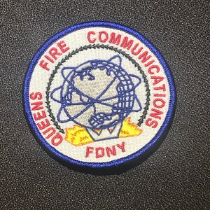 Könnte beinhalten: Ein weißer, runder Aufnäher mit blauer Umrandung. Der Aufnäher hat einen roten und schwarzen Kreis mit den Worten "FIRE COMMUNICATIONS" um einen weißen Globus mit einer roten und gelben Flamme darunter. Die Worte "QUEENS FDNY" sind um den äußeren Rand des Kreises geschrieben.