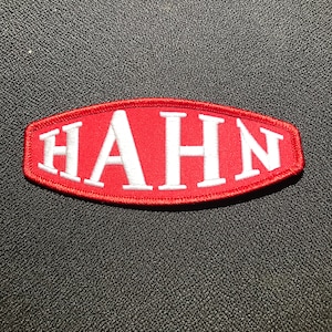 Könnte beinhalten: Roter ovaler Patch mit weiß gestickter Aufschrift "HAHN".