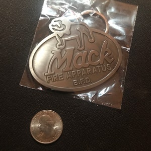Könnte beinhalten: Silberner Schlüsselanhänger mit einem Mack Fire Apparatus Logo. Das Logo zeigt einen stilisierten Hund und den Text "Mack Fire Apparatus B.F.D."