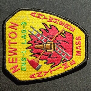 Puede incluir: Un parche bordado amarillo y rojo con el texto "NEWTON" y "ANYWHERE" en la parte superior e inferior, "ENG 1- LAD 3" a la izquierda y "MASS" a la derecha. El centro del parche presenta un fuego rojo y amarillo con una escalera y un hacha cruzadas.