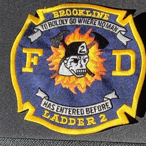 Puede incluir: Parche bordado con fondo azul y borde amarillo. El parche presenta el texto "BROOKLINE" y "TO BOLDLY GO WHERE NO MAN HAS ENTERED BEFORE LADDER 2". Una calavera con casco de bombero está en el centro, rodeada de llamas.