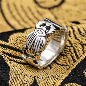 Op de afbeelding: Een zilveren ring met een gedetailleerd scarabee-keverontwerp. De ring heeft een brede band en ingewikkelde gravures, die de vleugels en het lichaam van de kever laten zien. De ring wordt getoond op een patroon van goud en zwarte stof.