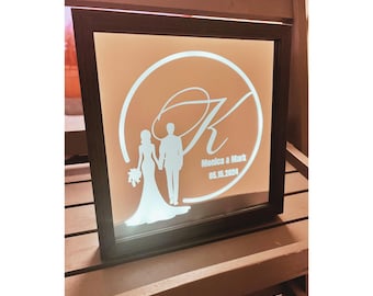May include: A white illuminated sign with a black frame. The sign features a stylized couple silhouette, a large letter "K" and the text "Monica & Mark 05.15.2024".