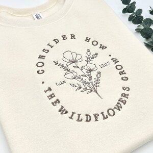 May include: A cream-colored sweatshirt with a brown embroidered design that says "Consider how the wildflowers grow" with a floral illustration and the text "Luke 12:27".