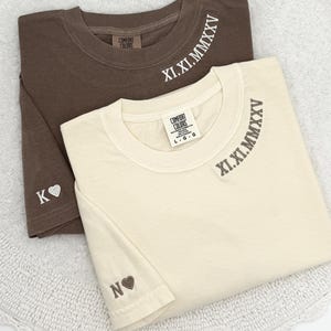 May include: Two folded t-shirts, one brown and one cream-colored, are displayed. The brown shirt has the Roman numeral "XI.XI.MMXXV" printed on the collar and the letter "K" with a heart on the sleeve. The cream shirt has the same Roman numeral and the letter "N" with a heart.
