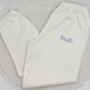 May include: White sweatpants with a drawstring waist and the word "bride" embroidered in light blue. The pants are folded and resting on a white, circular, textured surface.