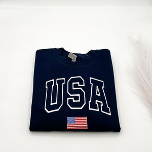 Puede incluir: Sudadera azul marino con la palabra "USA" en letras blancas. Debajo, una peque&ntilde;a bandera americana bordada. La sudadera est&aacute; doblada cuidadosamente sobre una superficie blanca.