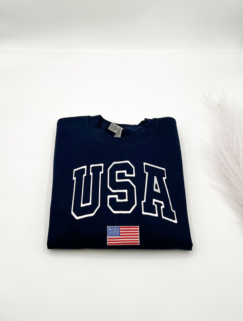 Puede incluir: Sudadera azul marino con la palabra "USA" en letras blancas. Debajo, una peque&ntilde;a bandera americana bordada. La sudadera est&aacute; doblada cuidadosamente sobre una superficie blanca.