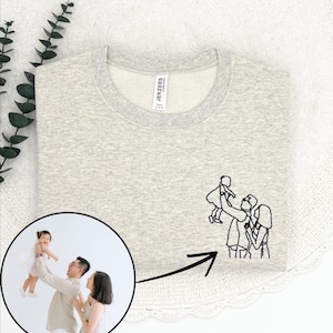 May include: A light gray sweatshirt with a crew neck and a small embroidered family portrait. The sweatshirt has a light gray collar and the brand name "JERZEES" is visible on a tag. A family photo is in a circle on the left.