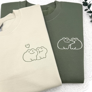 May include: Two sweatshirts, one beige and one olive green, are displayed. The beige sweatshirt features an embroidered design of two smiling frogs with a heart above them. The olive green sweatshirt has a similar design of two frogs. Both sweatshirts are made of a soft material.