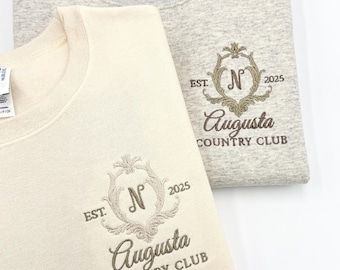 May include: Two folded sweatshirts, one cream and one light gray. Both feature an embroidered design with the text "Augusta Country Club", the letter "N", and the year "2025". The design is in a classic, elegant style.