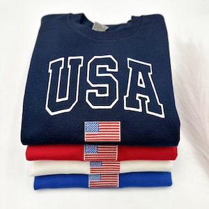 Puede incluir: Una pila de cuatro sudaderas en azul marino, rojo, blanco y azul. La sudadera azul marino superior tiene la palabra "USA" en letras blancas. Cada sudadera tiene una peque&ntilde;a bandera americana bordada.