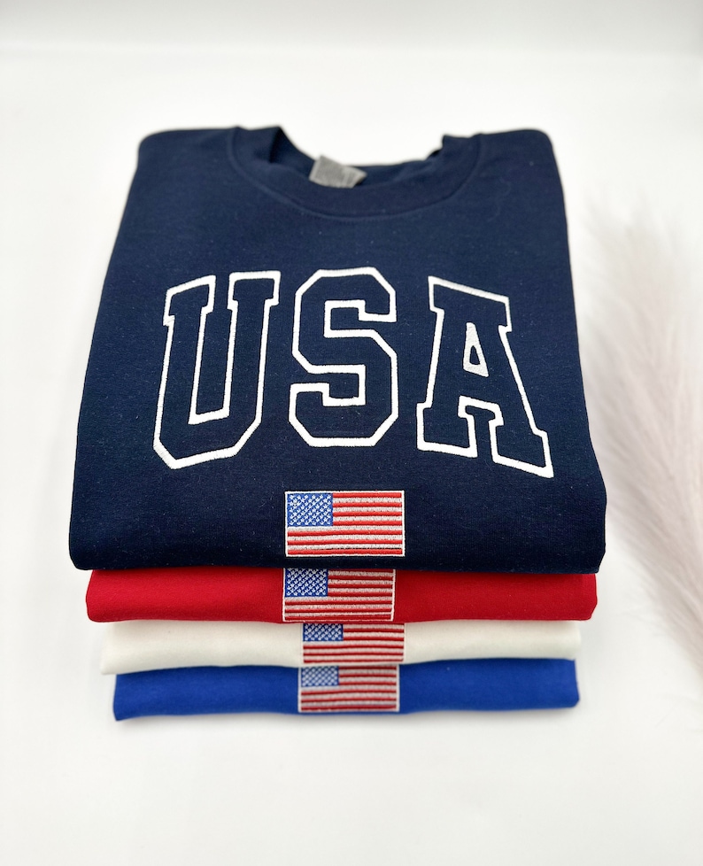 Puede incluir: Una pila de cuatro sudaderas en azul marino, rojo, blanco y azul. La sudadera azul marino superior tiene la palabra "USA" en letras blancas. Cada sudadera tiene una peque&ntilde;a bandera americana bordada.