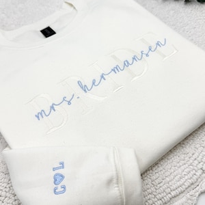 May include: Cream-colored sweatshirt with the word "BRIDE" and "mrs. hermansen" embroidered in light blue. The sleeve cuff displays the initials "COL" and a heart symbol, also in light blue. The sweatshirt is folded on a textured surface.
