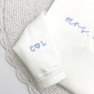 May include: Ivory-colored sweatshirt with light blue embroidered details. The sleeve features "C L" with a heart symbol. The word "Mrs." is embroidered on the front. The sweatshirt is on a white crocheted surface.