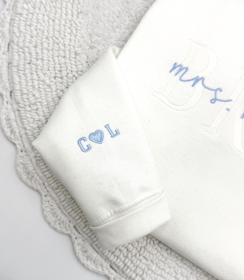 May include: Ivory-colored sweatshirt with light blue embroidered details. The sleeve features "C L" with a heart symbol. The word "Mrs." is embroidered on the front. The sweatshirt is on a white crocheted surface.