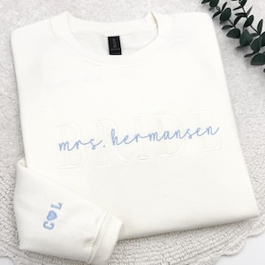 May include: Cream-colored sweatshirt with the embroidered text "BRIDE" and "mrs. hermansen" in light blue. The left sleeve cuff features the initials "C L" with a heart symbol. The sweatshirt is folded on a white surface.