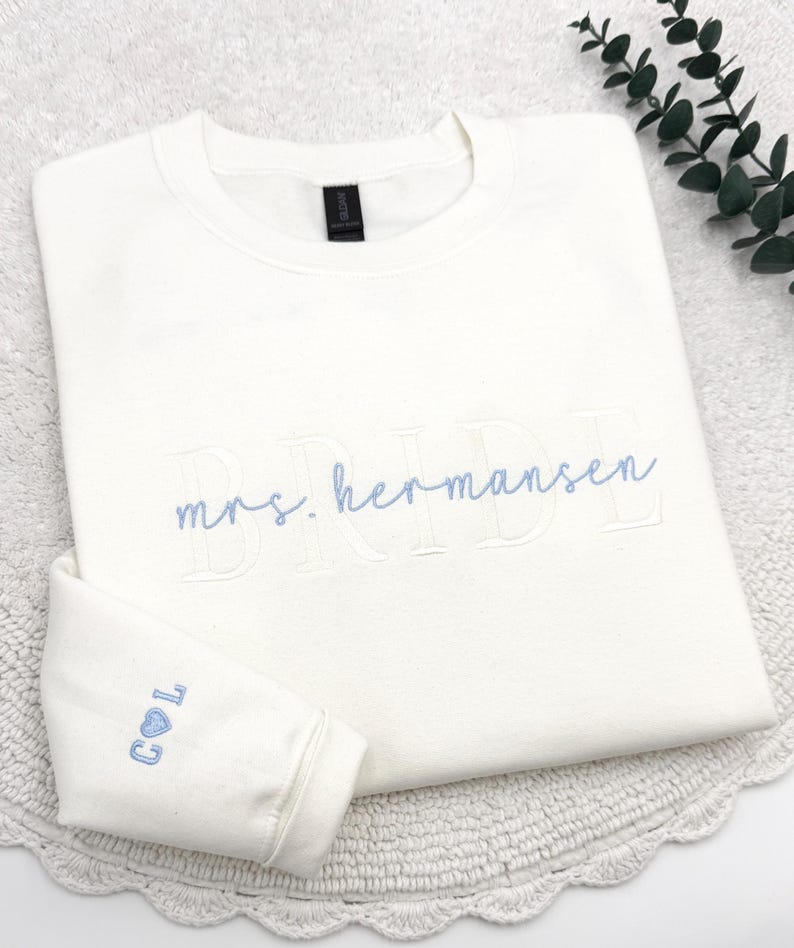 May include: Cream-colored sweatshirt with the embroidered text "BRIDE" and "mrs. hermansen" in light blue. The left sleeve cuff features the initials "C L" with a heart symbol. The sweatshirt is folded on a white surface.