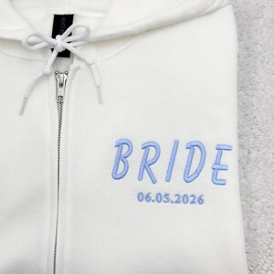 May include: Cream-colored zip-up hoodie with the word "BRIDE" embroidered in light blue, along with the date "06.05.2026". The hoodie features a drawstring hood and a silver zipper.