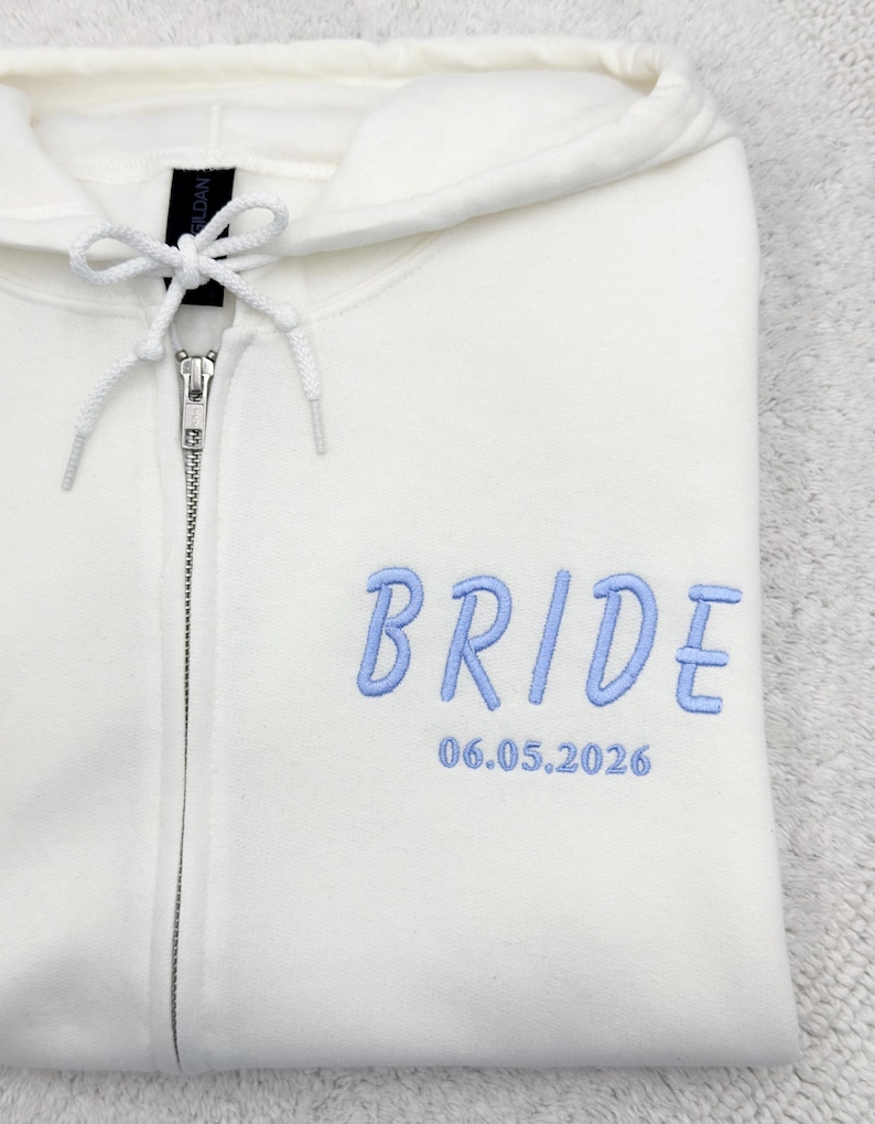 May include: Cream-colored zip-up hoodie with the word "BRIDE" embroidered in light blue, along with the date "06.05.2026". The hoodie features a drawstring hood and a silver zipper.