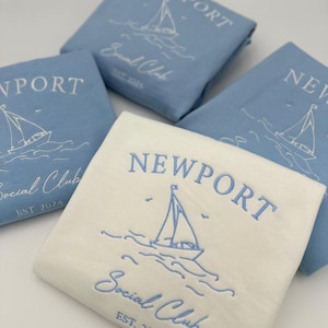 May include: Three folded white and light blue t-shirts with the text "NEWPORT Social Club EST. 2024" and a sailboat graphic.