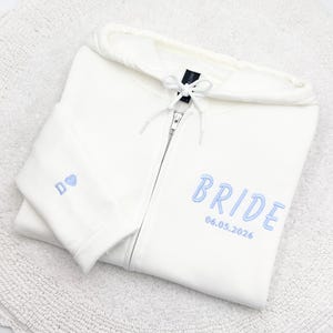 May include: Cream-colored zip-up hoodie with the word "BRIDE" embroidered in light blue, along with the date "06.05.2026". The left sleeve features the letter "D" and a blue heart. The hoodie is folded on a white surface.