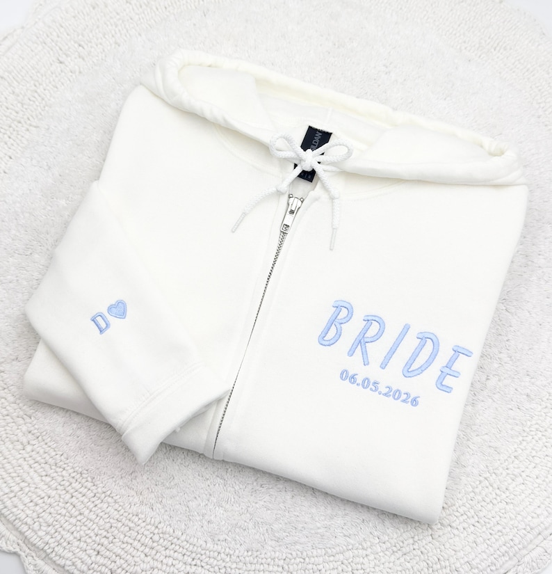 May include: Cream-colored zip-up hoodie with the word "BRIDE" embroidered in light blue, along with the date "06.05.2026". The left sleeve features the letter "D" and a blue heart. The hoodie is folded on a white surface.