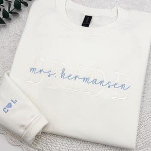 May include: Cream-colored sweatshirt with the embroidered text "mrs. Hermansen BRIDE" in light blue. The left sleeve cuff features the initials "C L" with a heart symbol. The sweatshirt is folded on a white surface with a decorative edge.