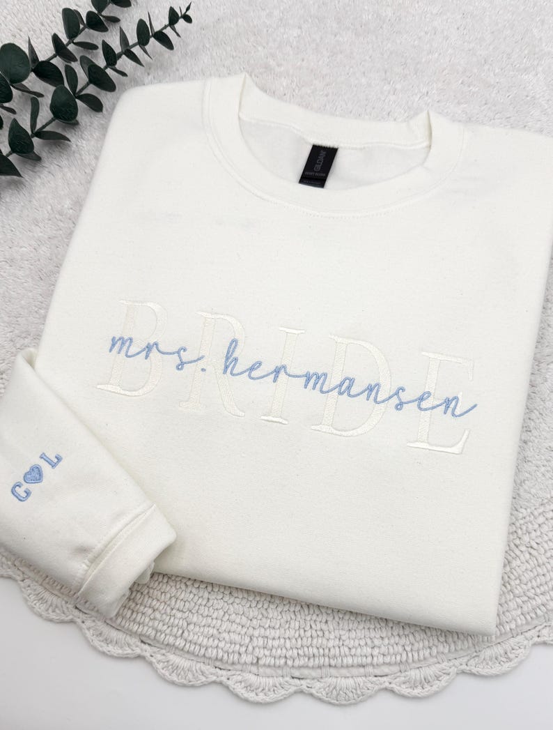 May include: Cream-colored sweatshirt with the embroidered text "mrs. Hermansen BRIDE" in light blue. The left sleeve cuff features the initials "C L" with a heart symbol. The sweatshirt is folded on a white surface with a decorative edge.