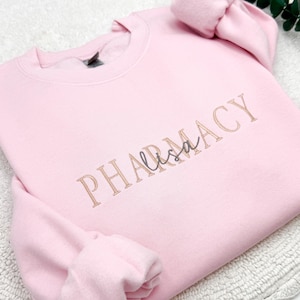 May include: A light pink crewneck sweatshirt with the word "PHARMACY" embroidered in a light brown color. The name "Lisa" is embroidered in a cursive font in the center of the word. The sweatshirt is made of a soft material.