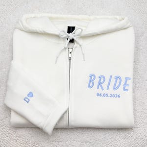 May include: Ivory zip-up hoodie with light blue embroidered text that reads "BRIDE" and the date "06.05.2026". The left sleeve has a small embroidered design. The hoodie has a drawstring hood and a silver zipper.