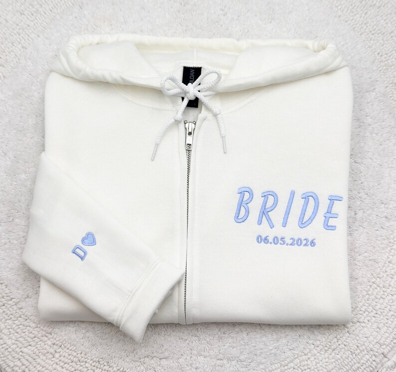 May include: Ivory zip-up hoodie with light blue embroidered text that reads "BRIDE" and the date "06.05.2026". The left sleeve has a small embroidered design. The hoodie has a drawstring hood and a silver zipper.
