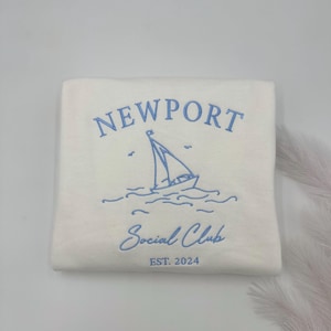 May include: A white sweatshirt with blue embroidered text that reads "NEWPORT Social Club EST. 2024". The design also includes a sailboat with waves.
