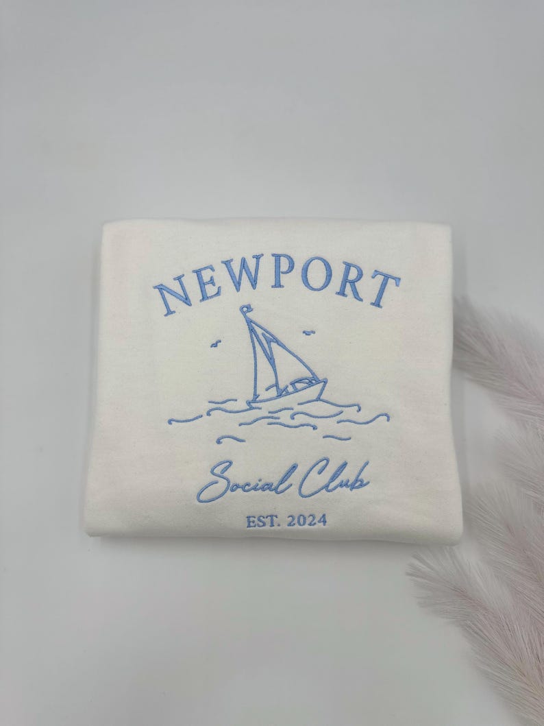 May include: A white sweatshirt with blue embroidered text that reads "NEWPORT Social Club EST. 2024". The design also includes a sailboat with waves.